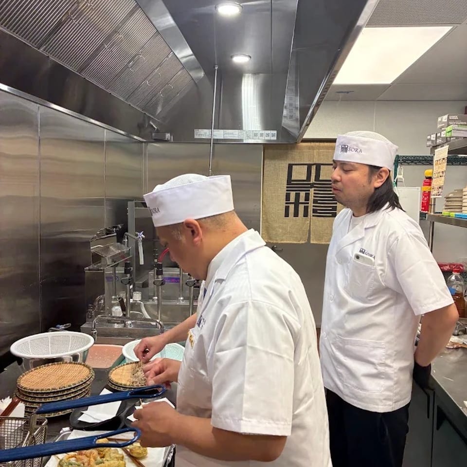 Two chefs prepare Japanese cuisine in a stainless-steel commercial kitchen, with tempura baskets and cooking equipment visible.