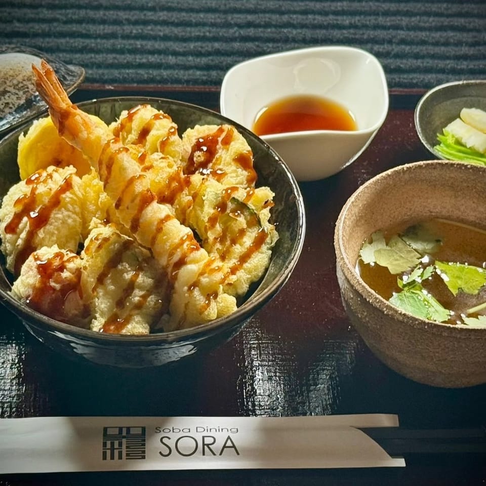 Japanese tempura rice bowl drizzled with sauce, accompanied by dipping sauce, soup, pickles, and Soba Dining SORA chopsticks.