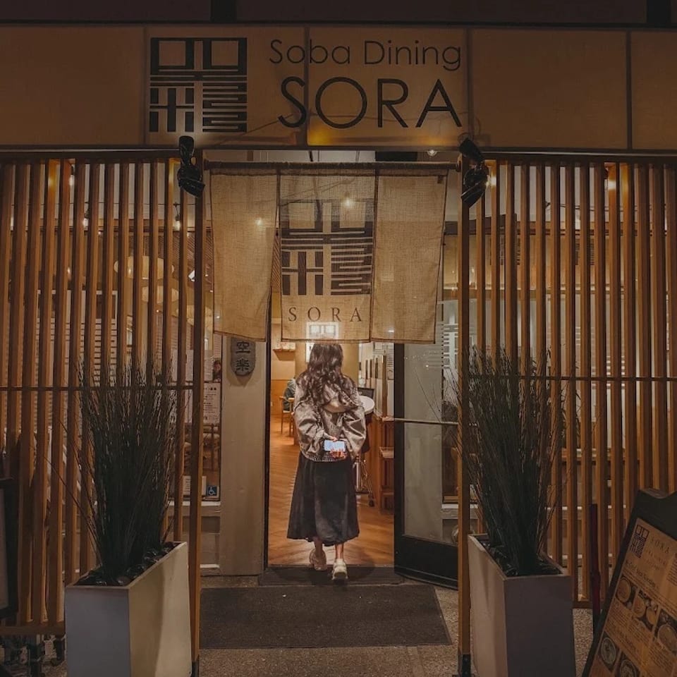 Soba Dining SORA restaurant entrance with Japanese-inspired wood screens, noren curtain, potted grasses, and a person entering.