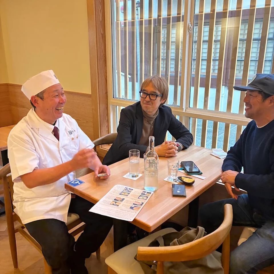 Chef and two guests chat around a restaurant table beside large windows, with menus, drinks, and smartphones on the table.