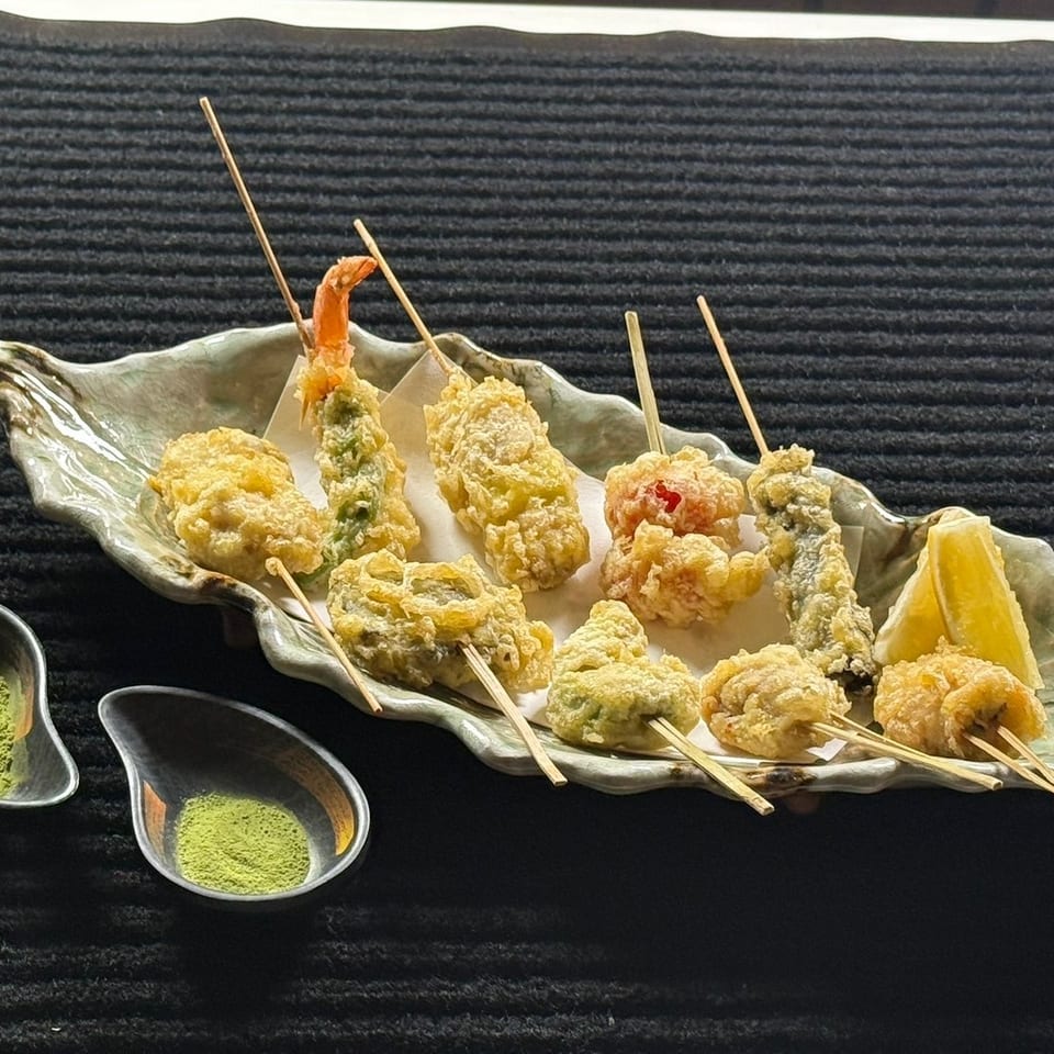Assorted tempura skewers, including shrimp and vegetables, served on a leaf-shaped platter with lemon and green tea dipping powder.
