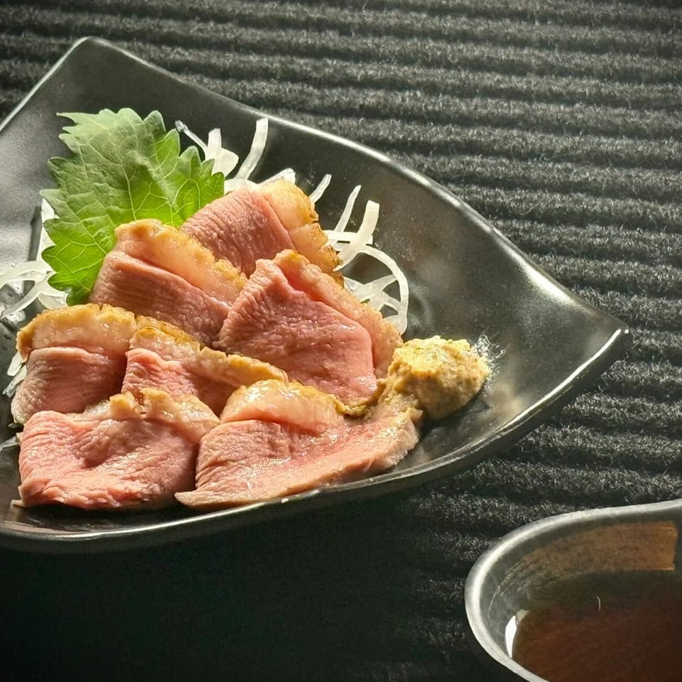 Sliced seared meat served on shredded daikon with shiso leaves, mustard, and dipping sauce on a black plate.