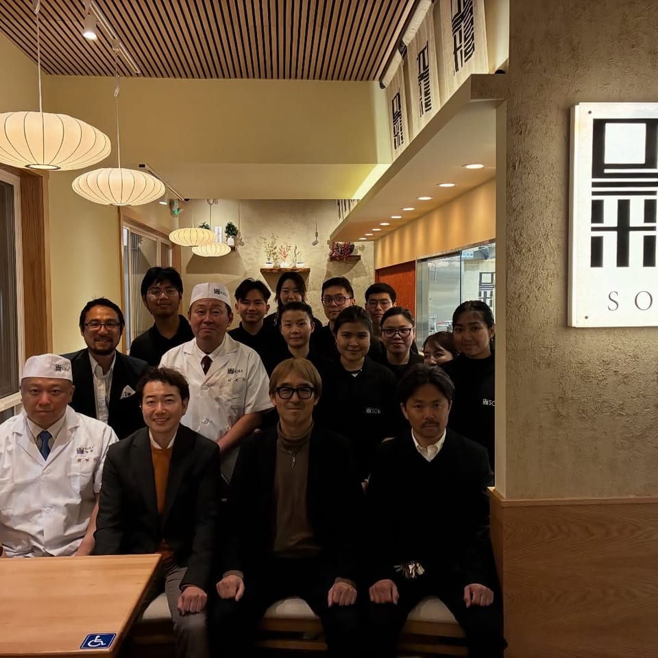 Restaurant staff pose beneath hanging lanterns beside a wall sign reading SORA.
