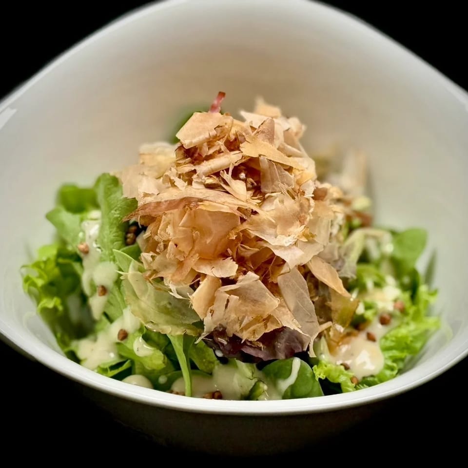 Fresh green salad topped with delicate bonito flakes, sesame seeds, and creamy dressing in a white bowl.