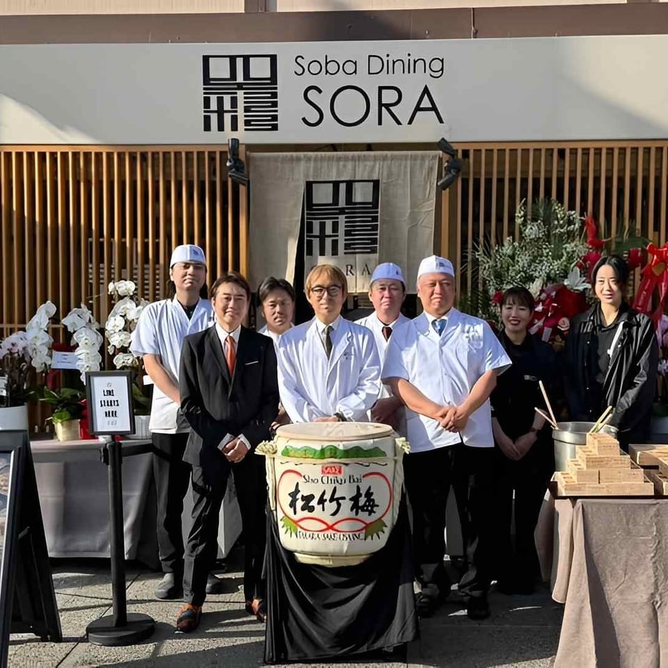 Soba Dining SORA staff pose outside the restaurant beside a ceremonial sake barrel, orchids, and stacked wooden gift boxes.