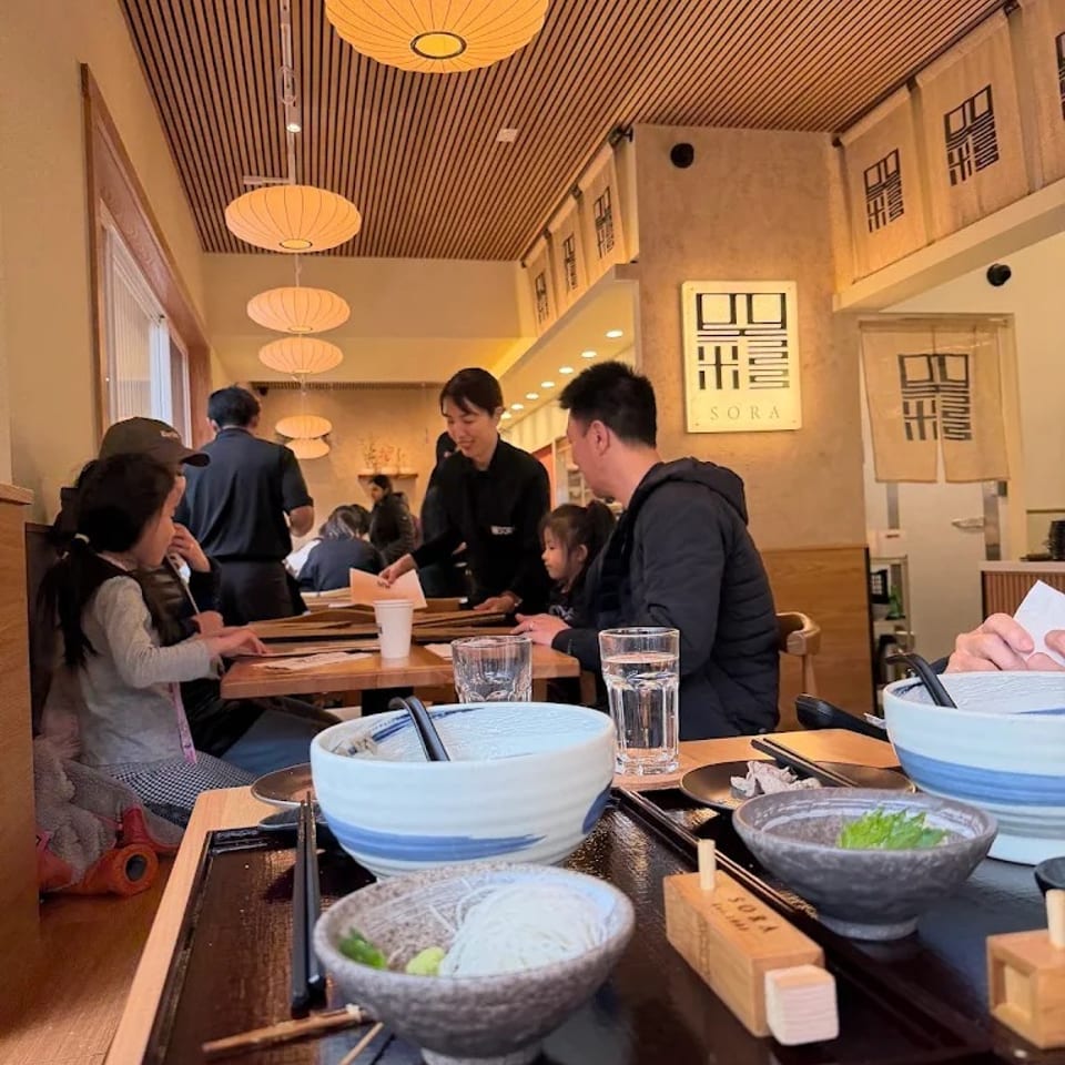 Busy Japanese restaurant interior with diners, bowls of noodles, drinks, warm pendant lighting, and SORA signage.