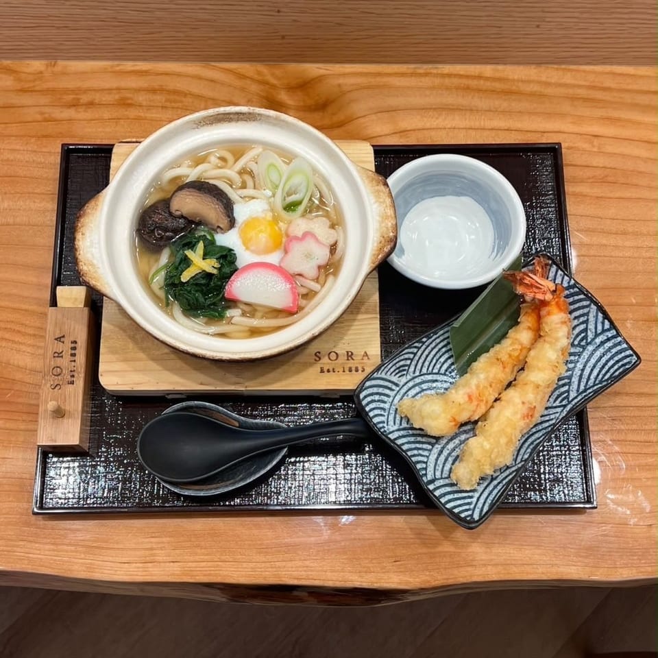 Japanese udon soup topped with vegetables and egg, served with shrimp tempura on a wooden restaurant tray.